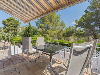 Terrasse extérieure avec carrelage en terre cuite, table en verre et chaises, sous un store rayé. Vues dégagées sur la verdure et les habitations lointaines.