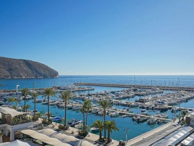 Vue panoramique sur une marina animée abritant de nombreux yachts et bateaux, bordée de palmiers et d'une mer turquoise. Un cap rocheux se profile à l'horizon.