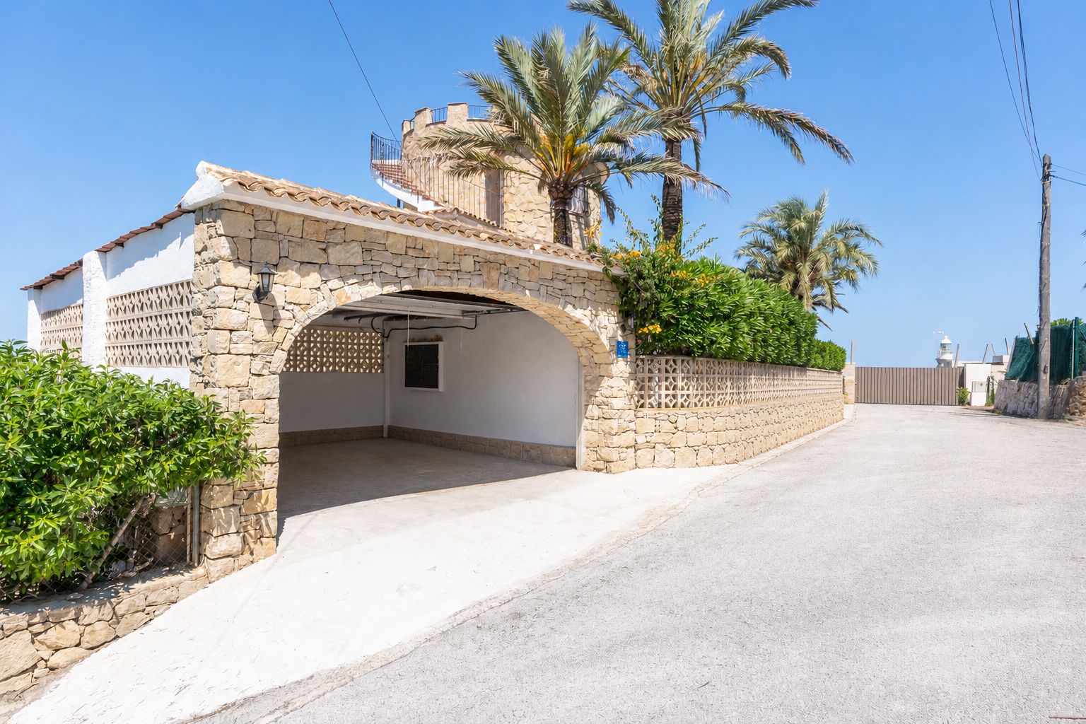 Abri de voiture voûté en pierre offrant un parking couvert, intégré à la villa de style méditerranéen avec escalier en colimaçon menant à une terrasse supérieure. Palmiers et verdure.