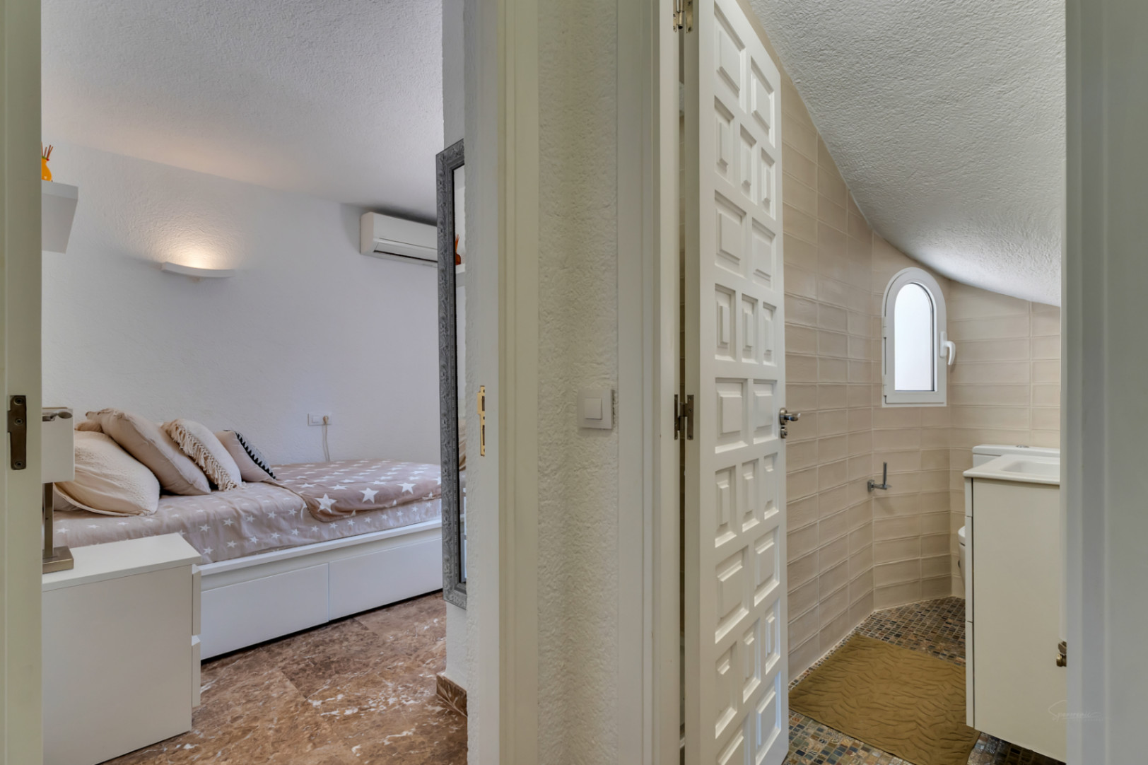 Aperçu d'une chambre avec sol effet marbre et climatisation, faisant face à une salle de bain aux murs carrelés beige, fenêtre arquée et sol en mosaïque avec meuble vasque.