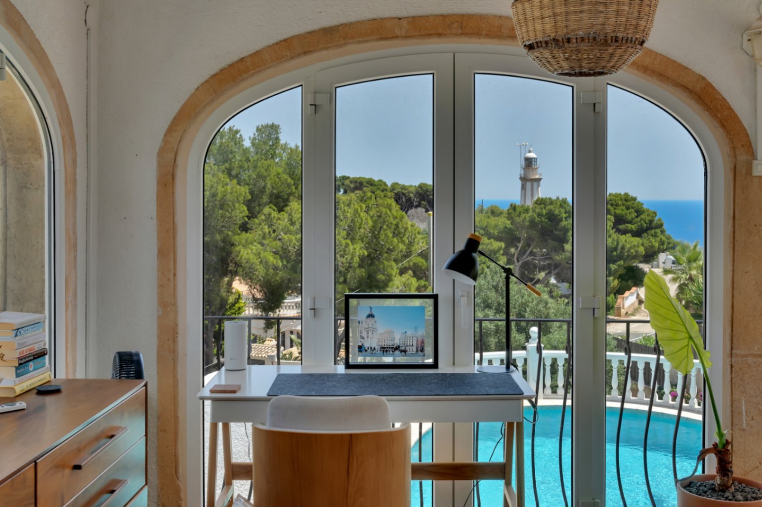 Bureau lumineux avec une fenêtre arquée offrant une vue panoramique sur la mer, le phare et la piscine.
