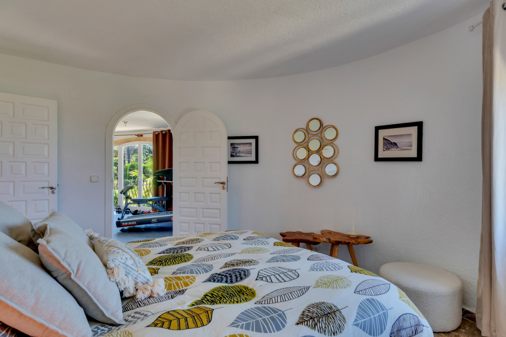 Chambre à coucher avec murs blancs texturés, lit design à motif de feuilles et porte cintrée ouvrant sur une pièce lumineuse avec équipement de sport.