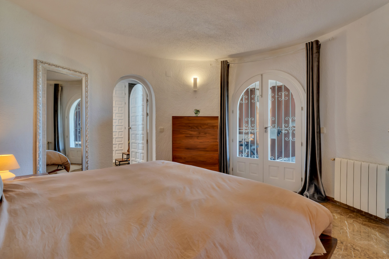 Chambre avec doubles portes cintrées blanches et grilles vers l'extérieur, sol effet marbre et commode en bois. Un grand miroir orné et des murs texturés enrichissent l'espace.