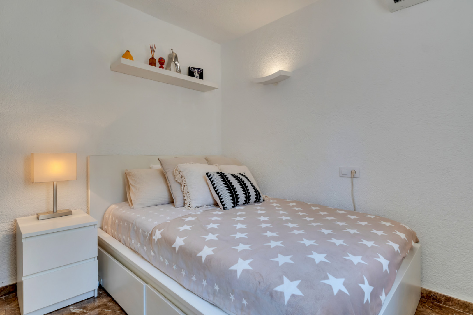 Chambre individuelle avec lit moderne, couette étoilée, table de chevet blanche et étagère murale décorée. Éclairage mural discret et climatisation.