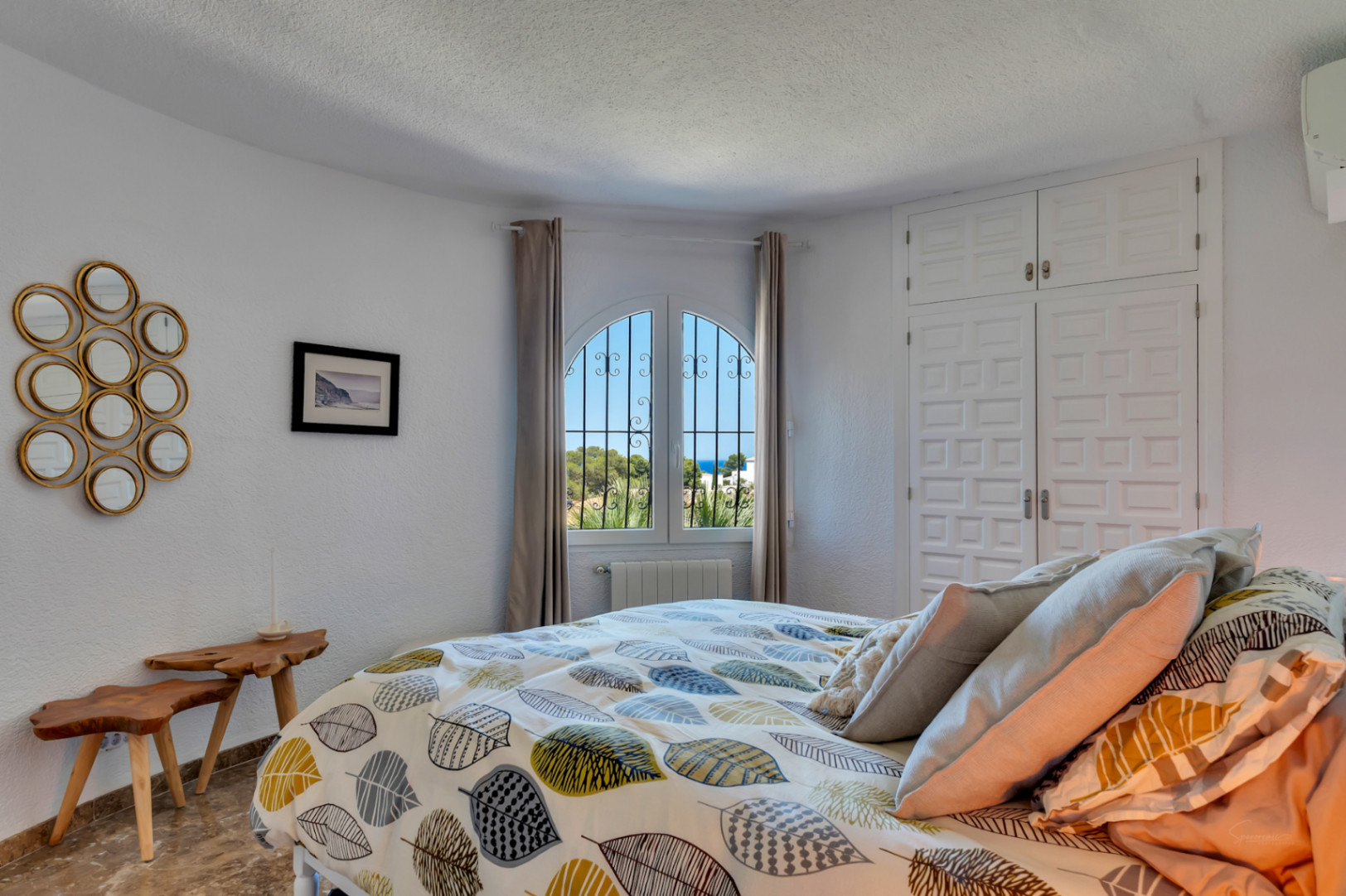 Chambre lumineuse avec fenêtre cintrée et barreaux en fer forgé, offrant une vue partielle sur la mer. Elle dispose d'armoires encastrées blanches et de tables de chevet en bois rustique.