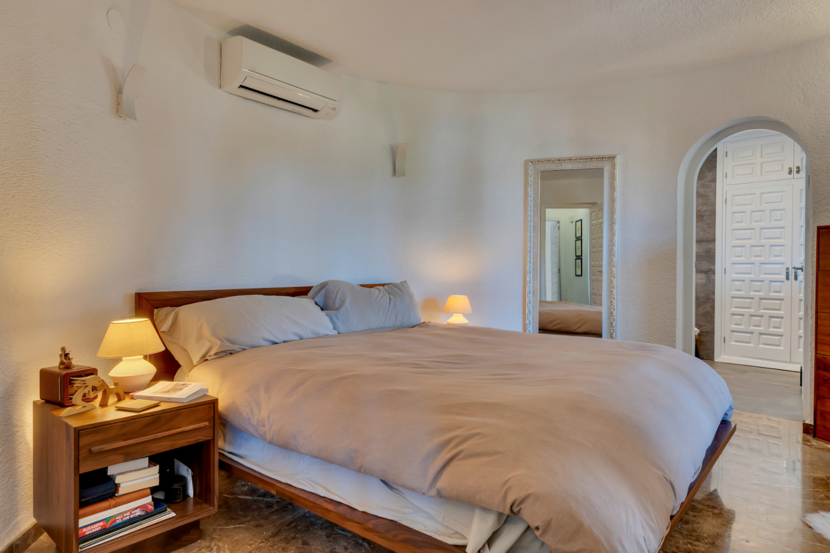 Chambre principale avec lit en bois, murs blancs texturés et climatisation. Un miroir orné et une arche mènent à une porte à motifs distinctifs.