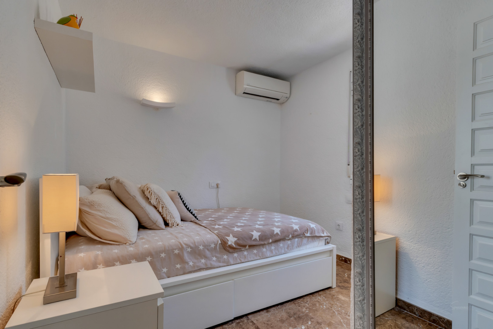 Chambre secondaire avec murs blancs texturés, un lit avec une couette à motif d'étoiles et une lampe de chevet moderne. Comprend une unité de climatisation et un sol carrelé.