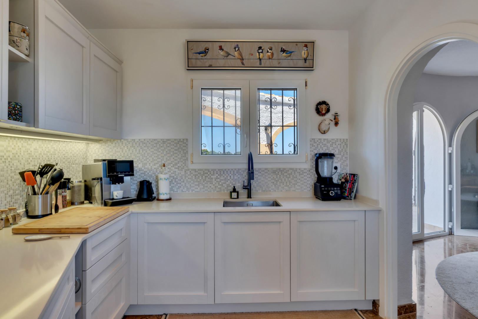 Cuisine moderne avec des armoires blanches, des comptoirs clairs et une crédence en mosaïque. Une fenêtre grillagée donne sur l'extérieur, et une arche mène à un espace lumineux adjacent.