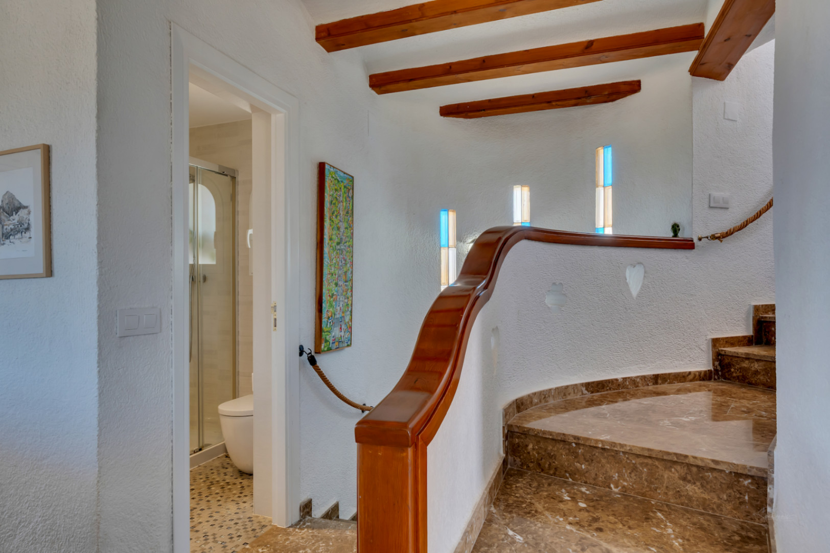 Escalier courbe en marbre brun avec rampe en bois, poutres apparentes et fenêtres à vitraux colorés. Accès à une salle de bain avec douche.
