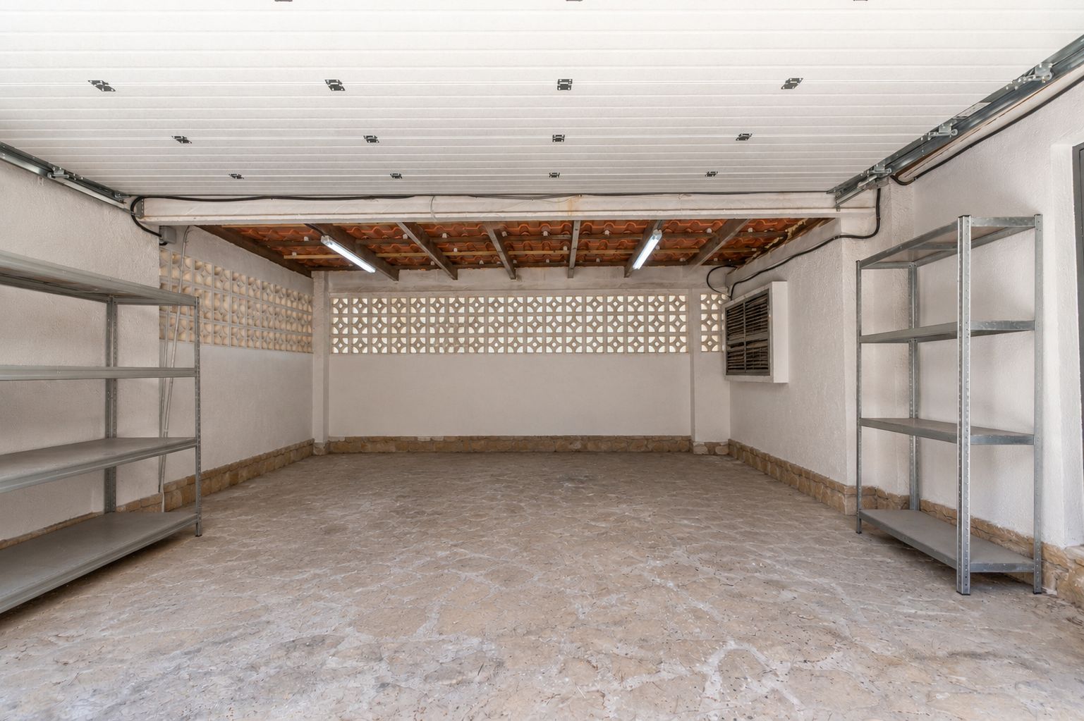 Grand garage avec sol en béton texturé, murs en stuc blanc et blocs de ventilation décoratifs. Avec tuiles en terre cuite apparentes, éclairage fluorescent et étagères métalliques.