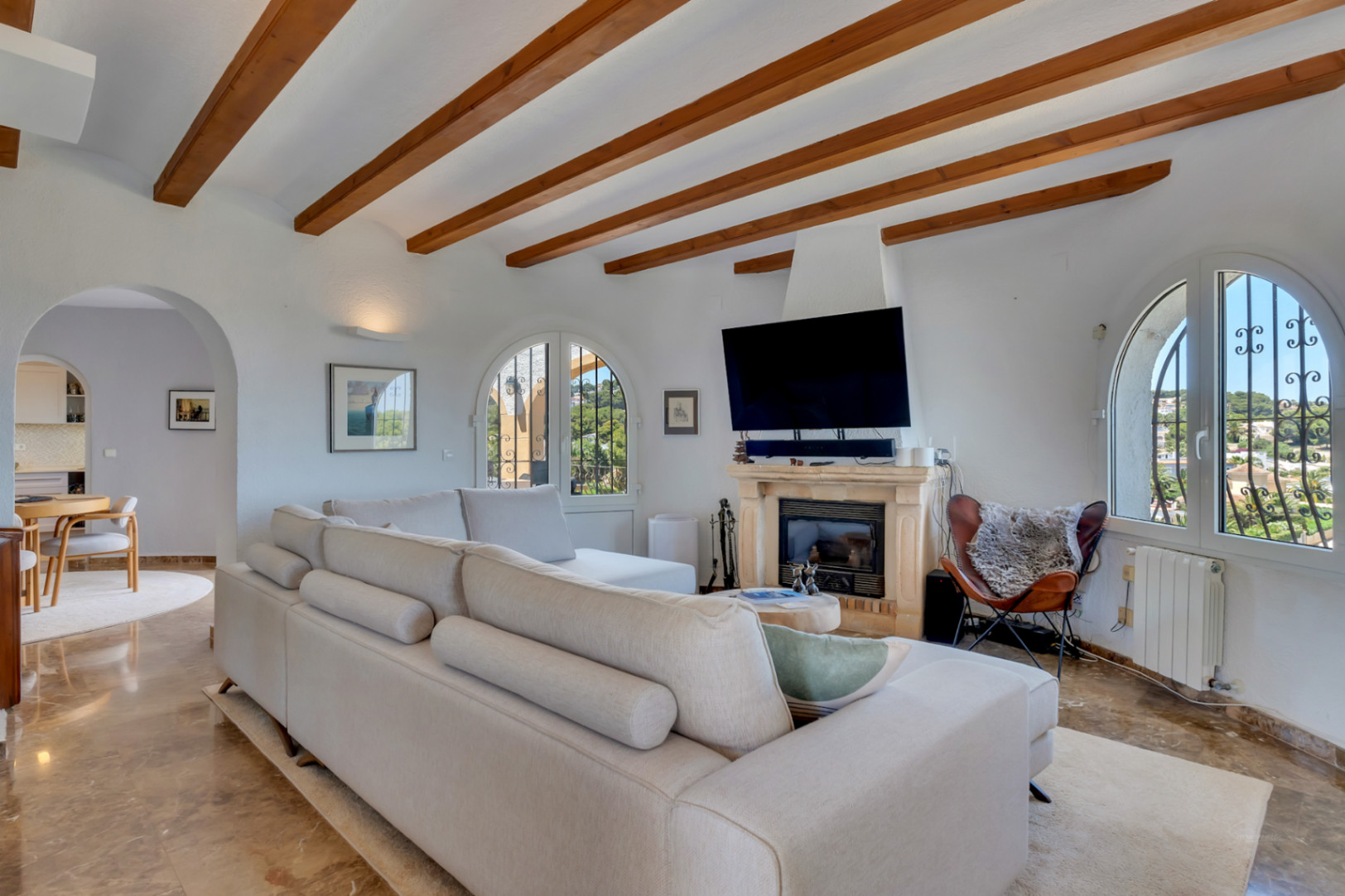 Grand salon avec poutres apparentes en bois, cheminée en pierre et fenêtres cintrées offrant des vues sur le paysage. Canapé modulaire clair et sol en marbre.