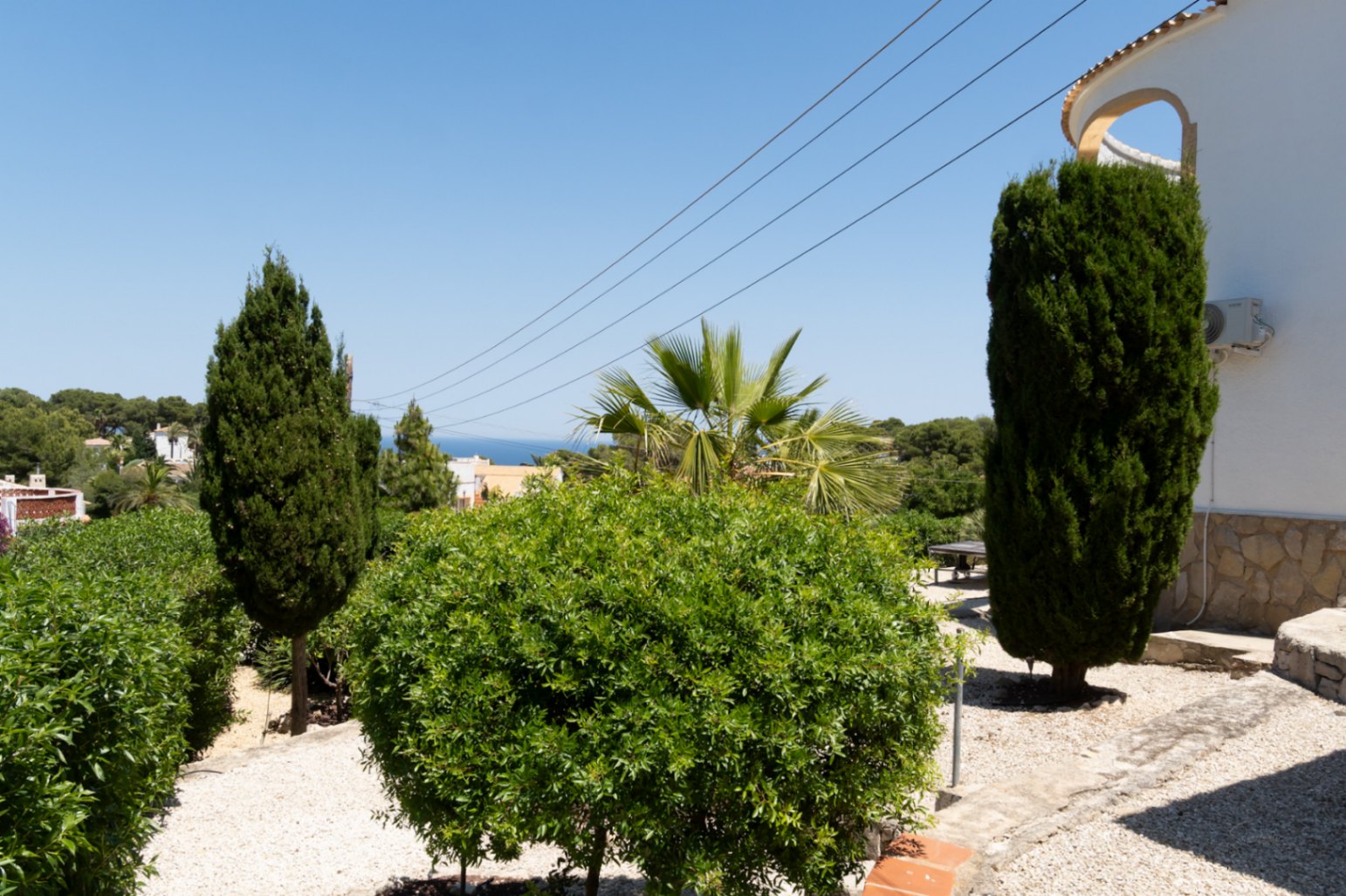 Jardin méditerranéen avec allée de gravier, végétation luxuriante et cyprès, offrant une vue lointaine sur la mer sous un ciel clair.