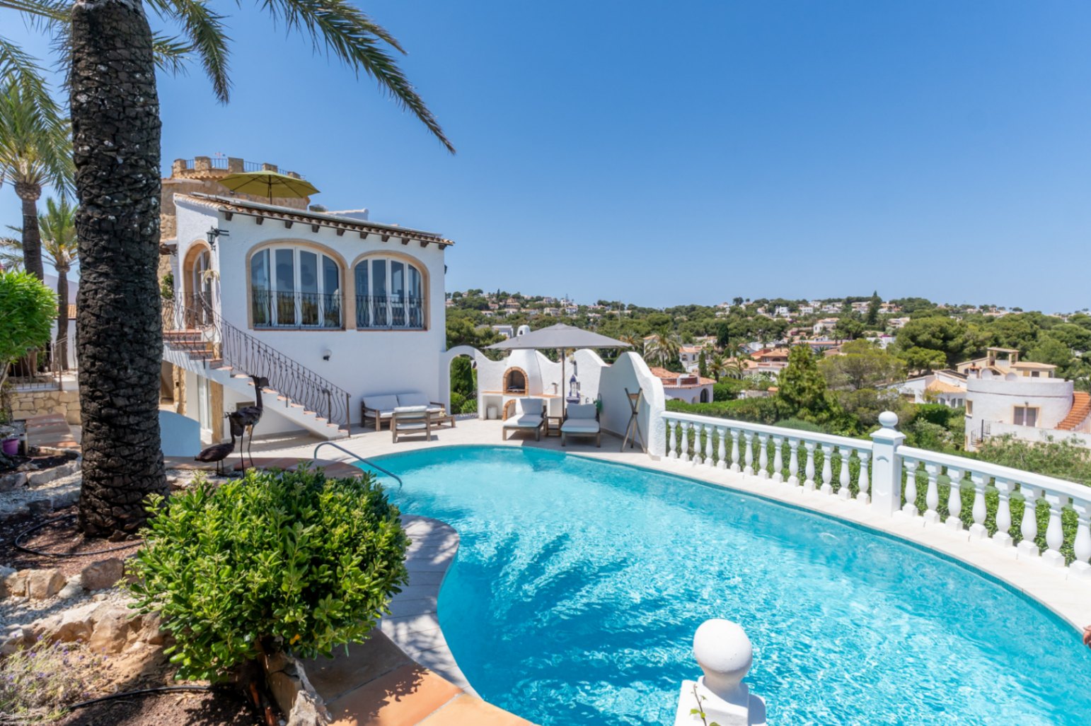 La piscine incurvée aux eaux claires se marie avec la villa méditerranéenne blanche, dotée de fenêtres cintrées et d'une cuisine extérieure avec chaises longues, entourée d'une végétation luxuriante.