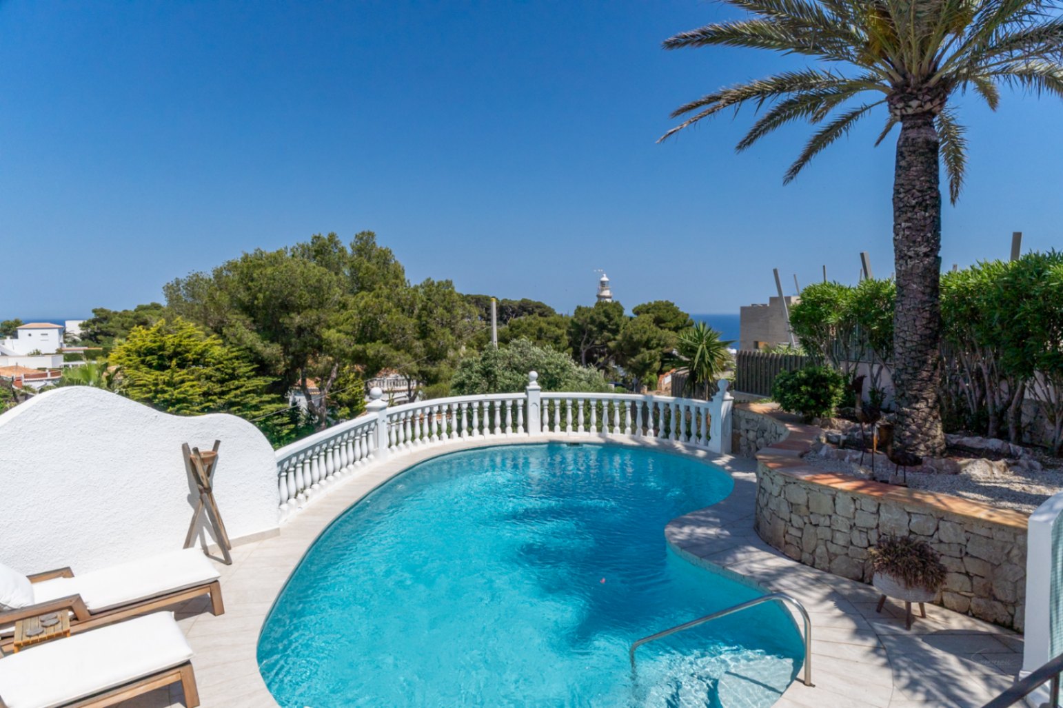 Piscine incurvée avec balustrade blanche, entourée de transats et d'un palmier. Vue lointaine sur la mer et un phare sous un ciel clair.