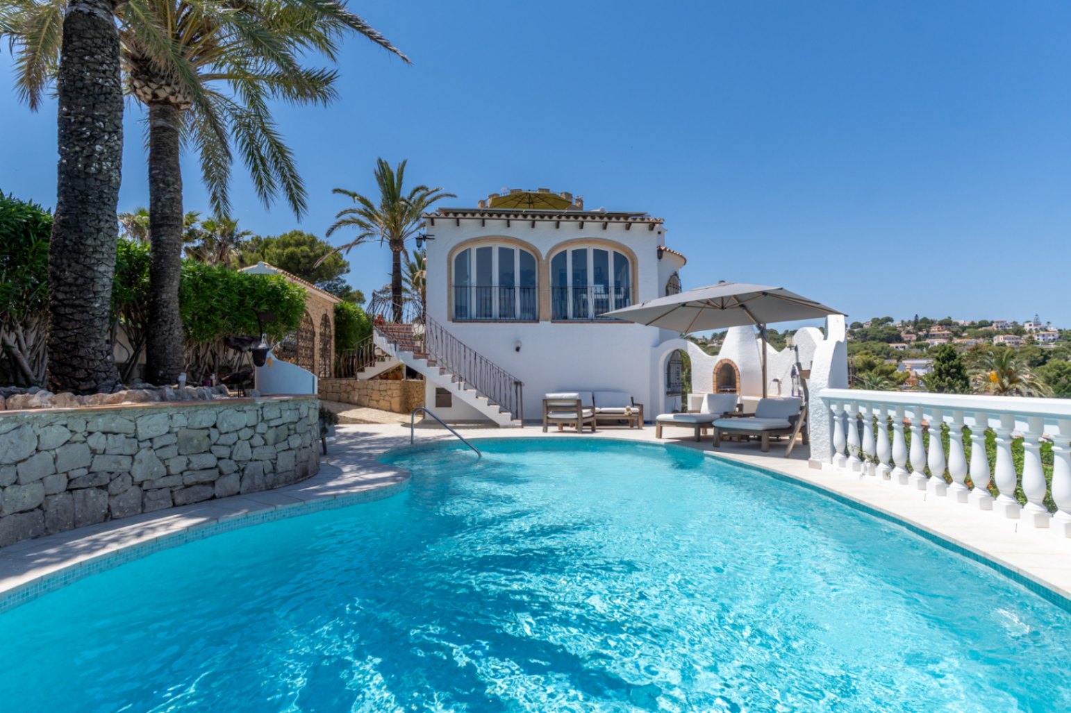 Piscine incurvée d'eau bleue avec espace solarium, barbecue extérieur et villa méditerranéenne blanche, entourée de palmiers et de verdure luxuriante.