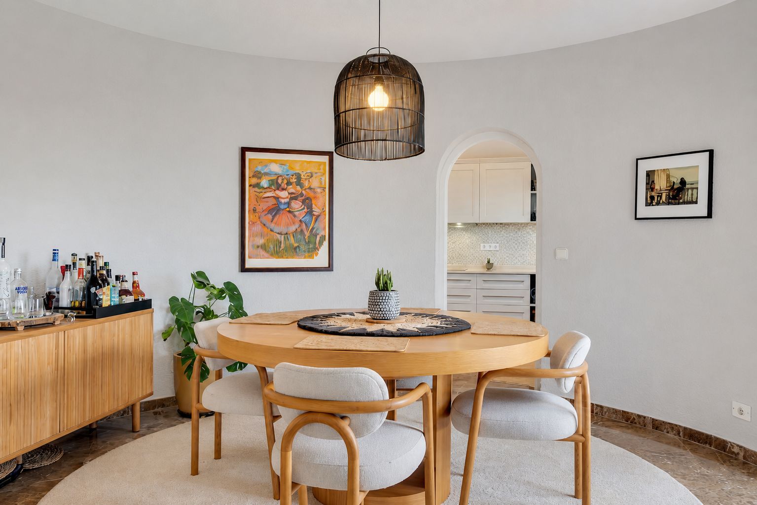 Salle à manger incurvée avec table ronde en bois, quatre chaises rembourrées et une suspension en osier. Une arche mène à la cuisine attenante.