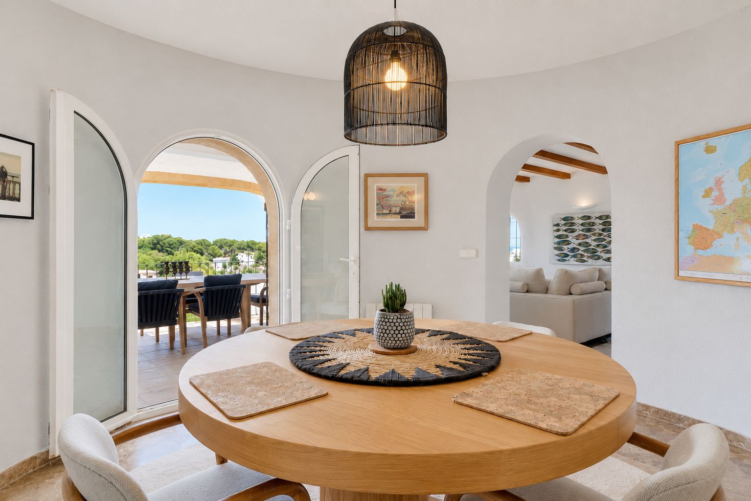 Salle à manger lumineuse avec table ronde en bois et suspension tressée. Des arches mènent à une terrasse extérieure verdoyante et à un salon confortable.