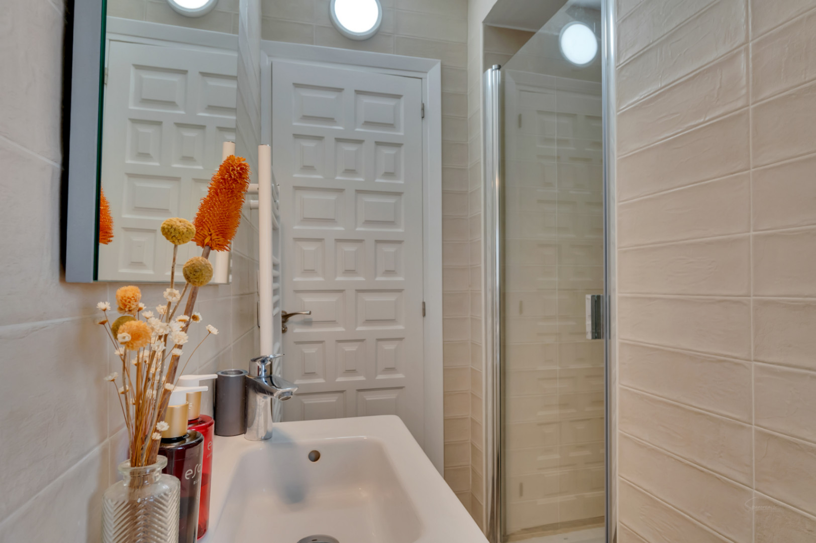 Salle de bain compacte avec lavabo blanc, robinetterie chromée et douche vitrée. Les murs sont carrelés de beige, porte à panneaux blancs.