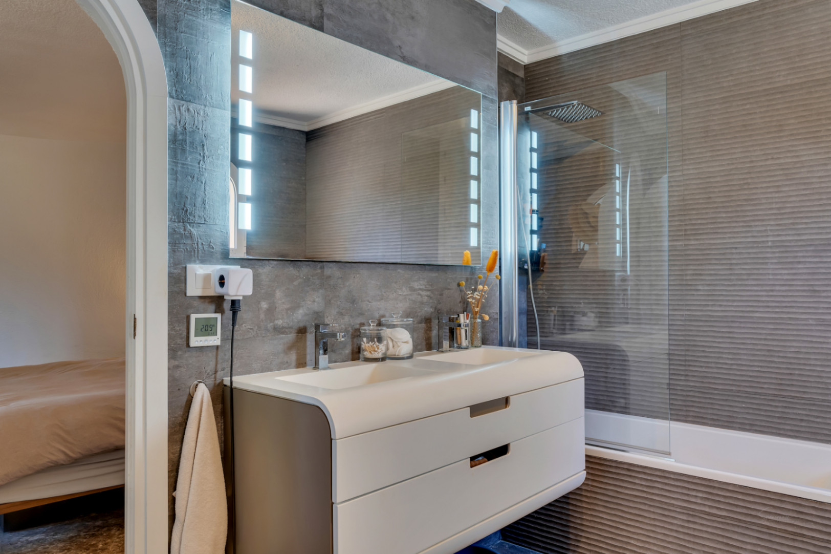 Salle de bain contemporaine avec double vasque, meuble incurvé blanc, miroir éclairé par LED et murs en carrelage gris foncé texturé. Une douche intégrée avec paroi en verre est présente.