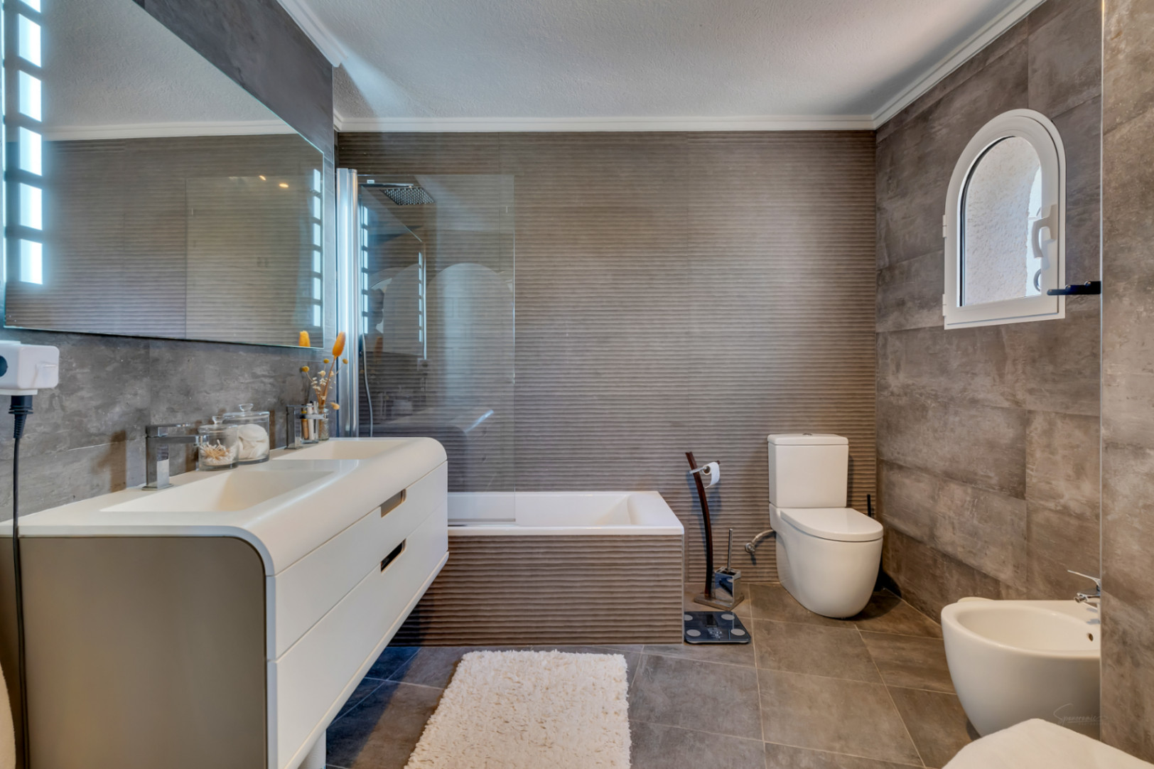 Salle de bain moderne avec carrelage gris texturé, double vasque flottante et miroir éclairé. Une fenêtre cintrée apporte du caractère au-dessus de la zone des toilettes.