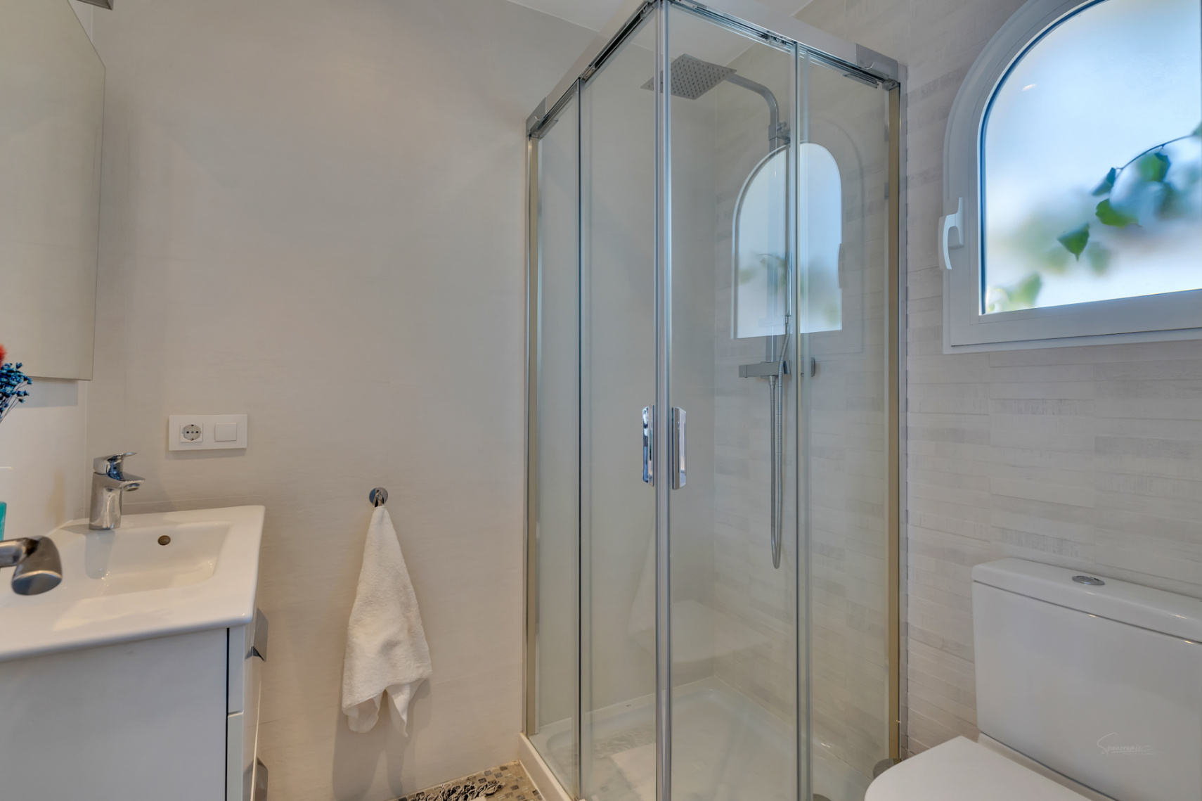 Salle de bain moderne avec une cabine de douche vitrée et pommeau de pluie. Une fenêtre arquée dépolie et un meuble-lavabo blanc complètent cet espace carrelé.