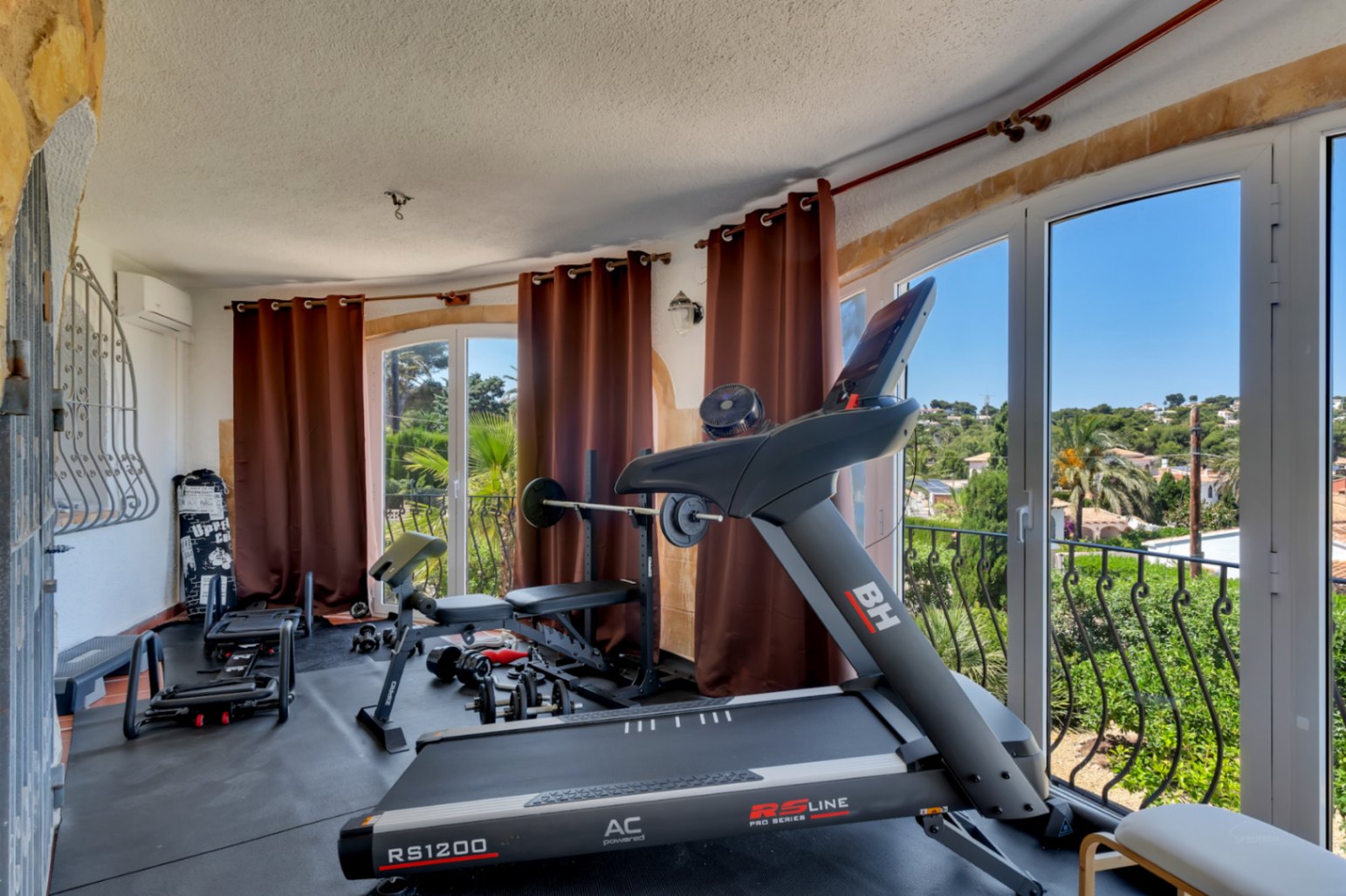 Salle de sport à domicile équipée d'un tapis de course et d'haltères, offrant des vues panoramiques sur la verdure et le paysage depuis ses grandes fenêtres.