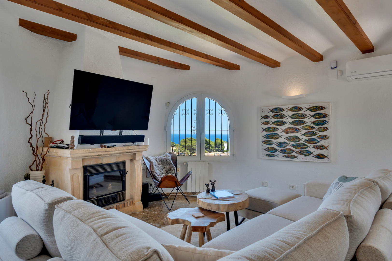 Salon avec poutres en bois apparentes et cheminée en pierre, offrant une vue sur la mer depuis une fenêtre arquée. Canapé modulaire et tables basses en bois naturel.