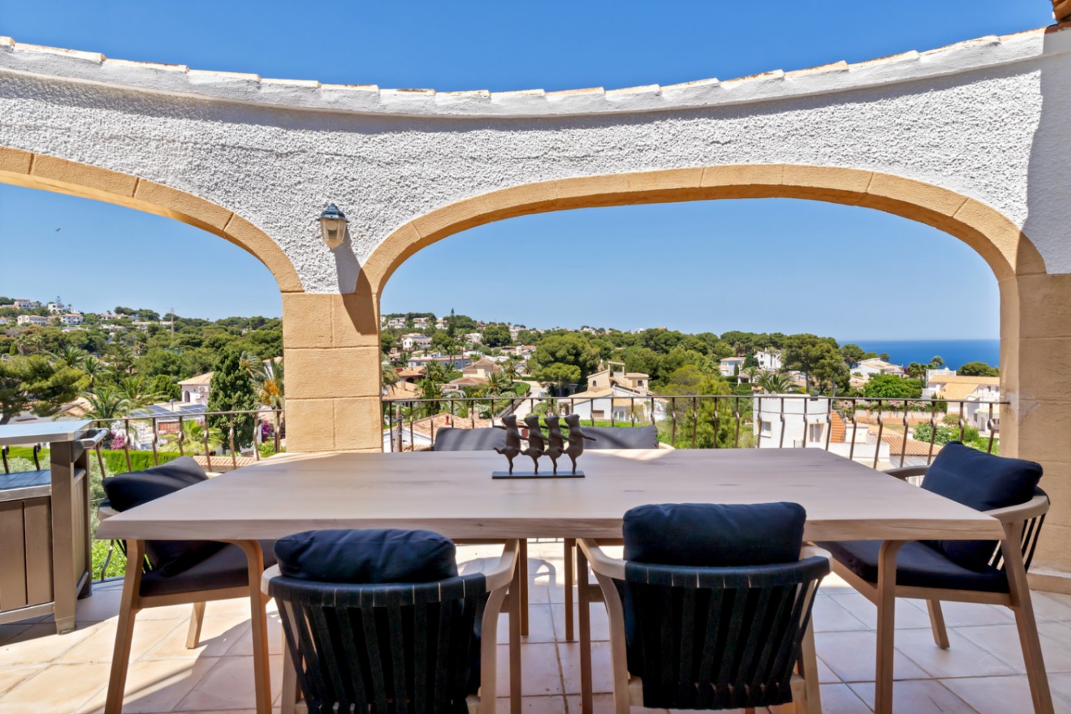Terrasse couverte avec une table à manger en bois clair et des chaises à coussins sombres, offrant des vues panoramiques sur la mer et la ville à travers des arches méditerranéennes.