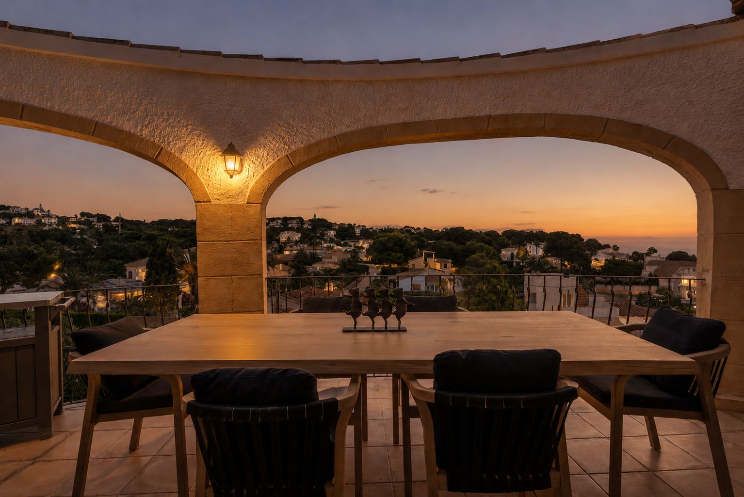 Terrasse élégante avec arches au crépuscule, dotée d'une table à manger en bois. Profitez des vues panoramiques sur la zone résidentielle illuminée et l'horizon marin.