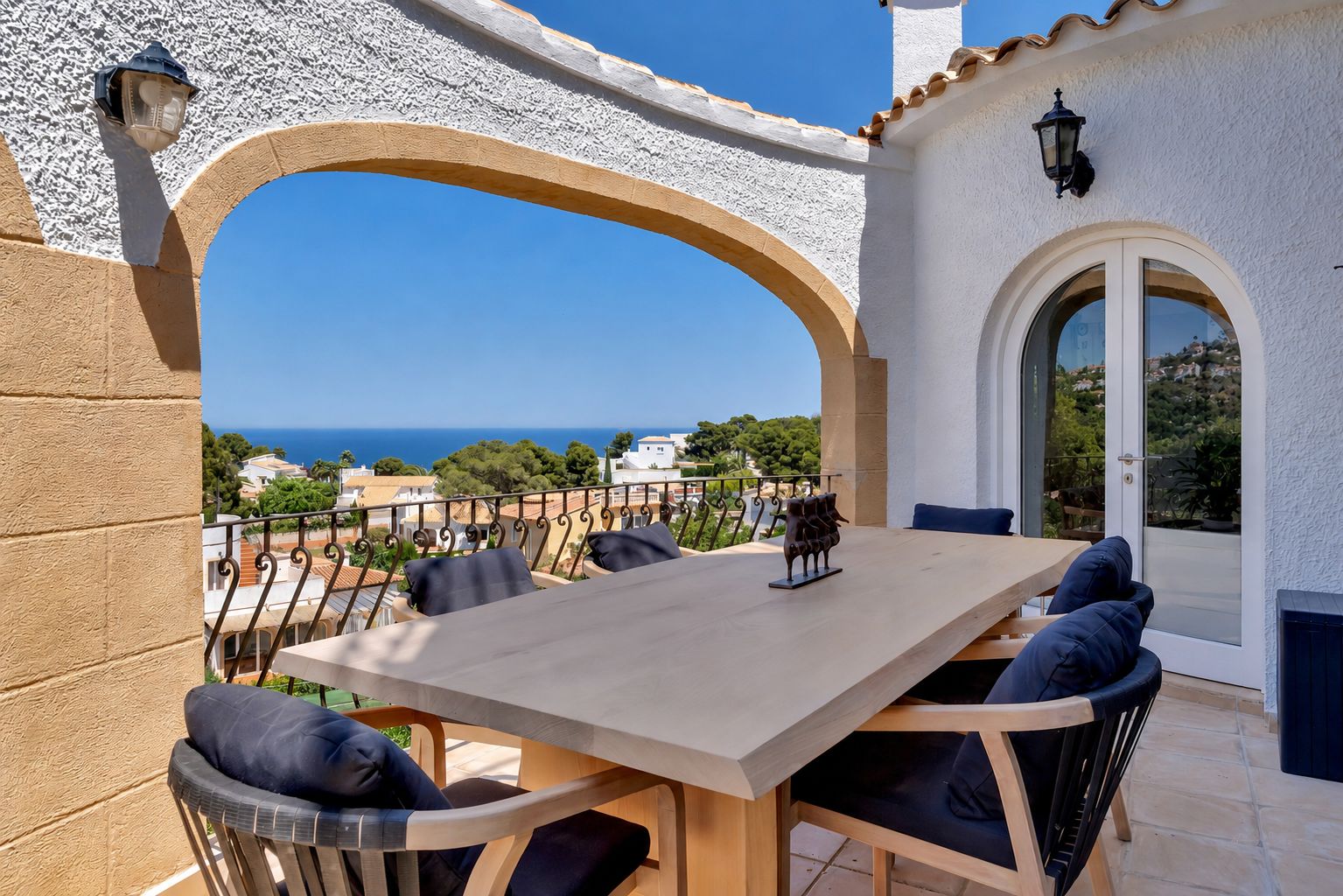 Terrasse repas extérieure avec une table en bois clair et des chaises aux coussins bleus. Une arche encadre des vues panoramiques sur la mer Méditerranée.