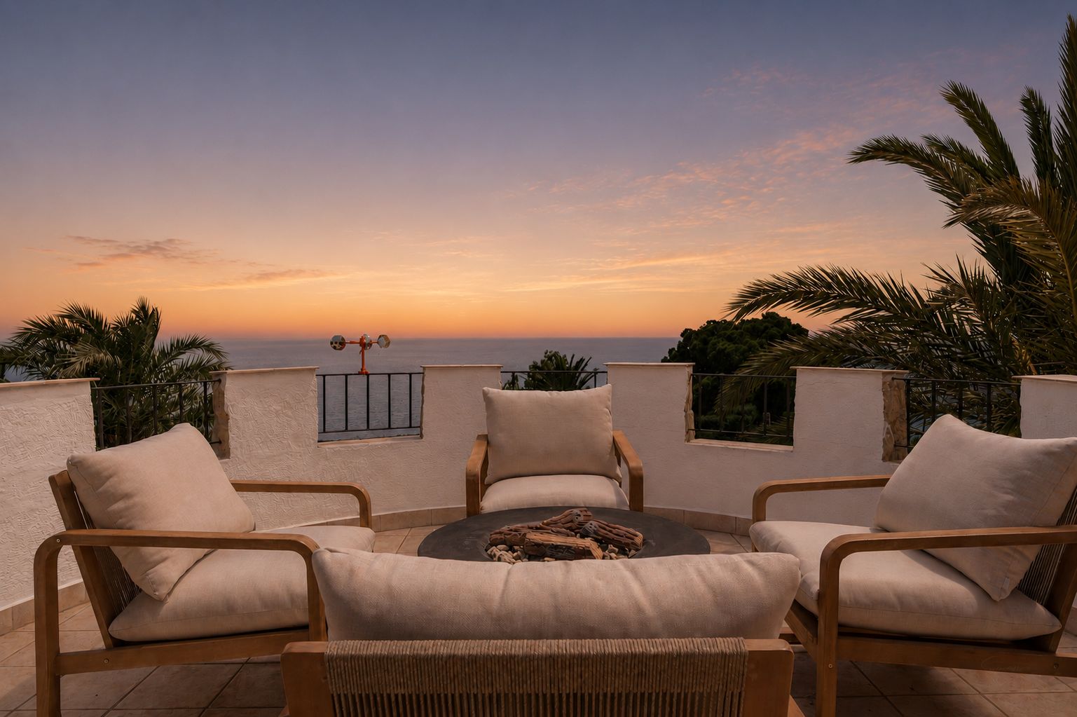 Terrasse sur le toit avec quatre chaises en bois et coussins clairs autour d'un foyer, offrant une vue panoramique sur la mer et des palmiers sous un ciel de coucher de soleil.