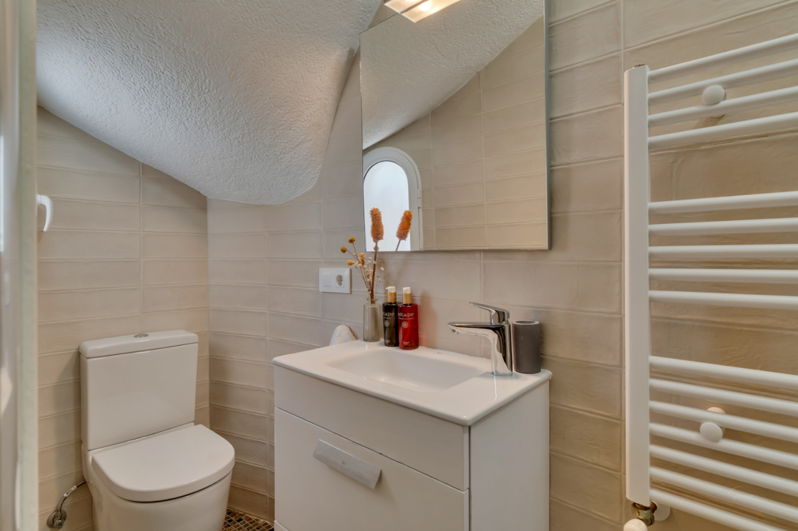 Toilettes fonctionnelles avec murs carrelés clairs, meuble-lavabo blanc et WC. Un miroir reflète une fenêtre arquée et un sèche-serviettes.