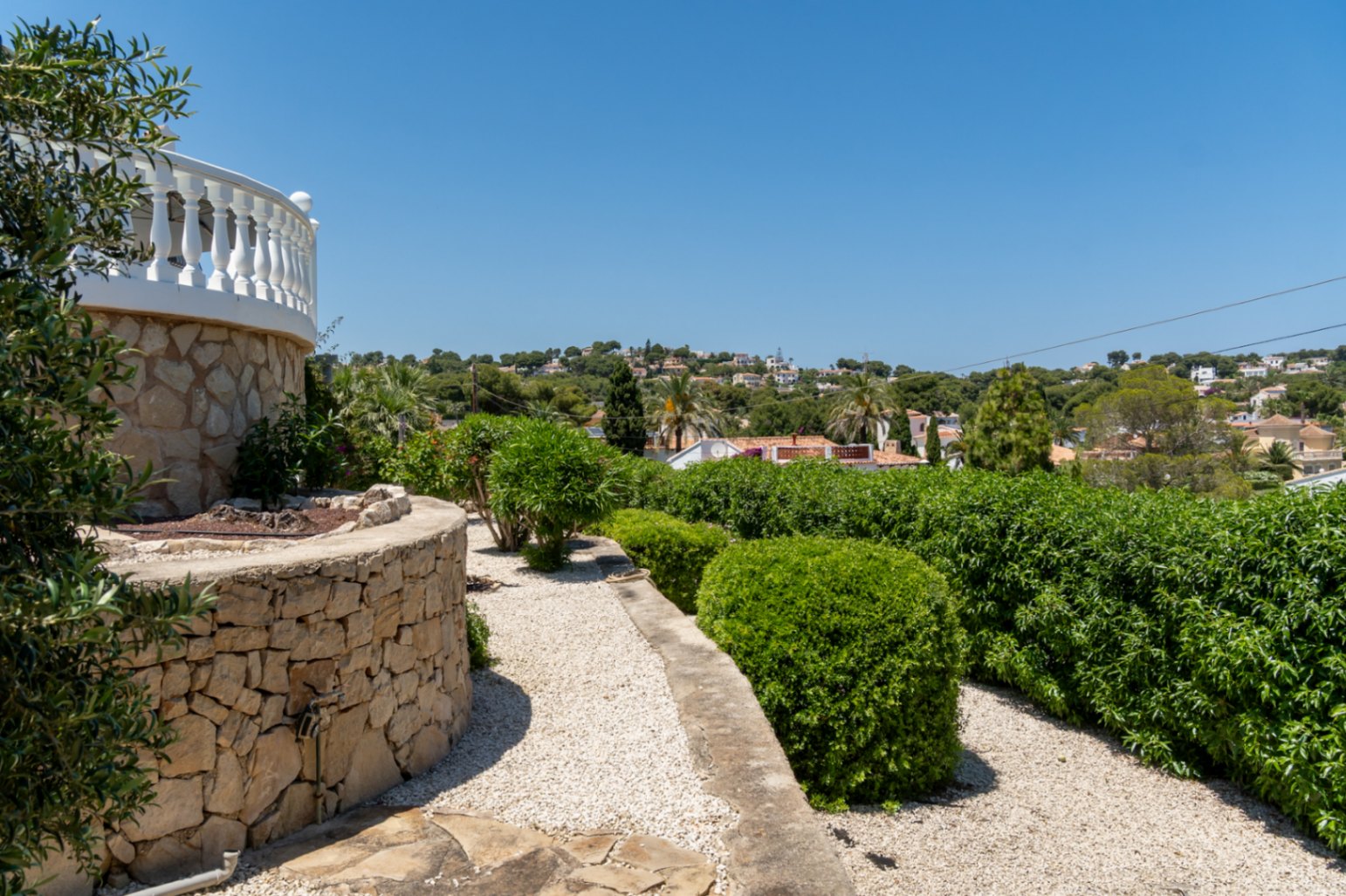 Un mur en pierre sèche incurvé soutient une balustrade blanche, encadrant un chemin de gravier bordé de haies taillées. Des maisons lointaines parsèment la colline verdoyante.