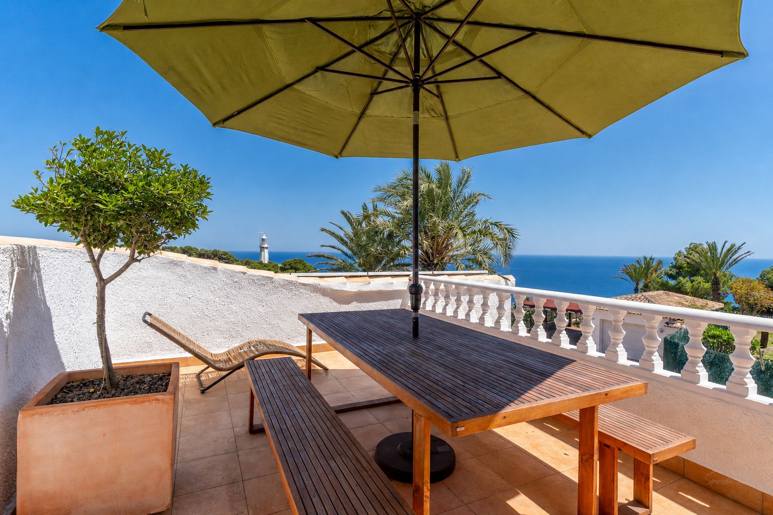 Vaste terrasse avec mobilier de repas en bois sous un parasol, offrant une vue panoramique sur la mer et un phare lointain. Sol carrelé et arbre en pot.