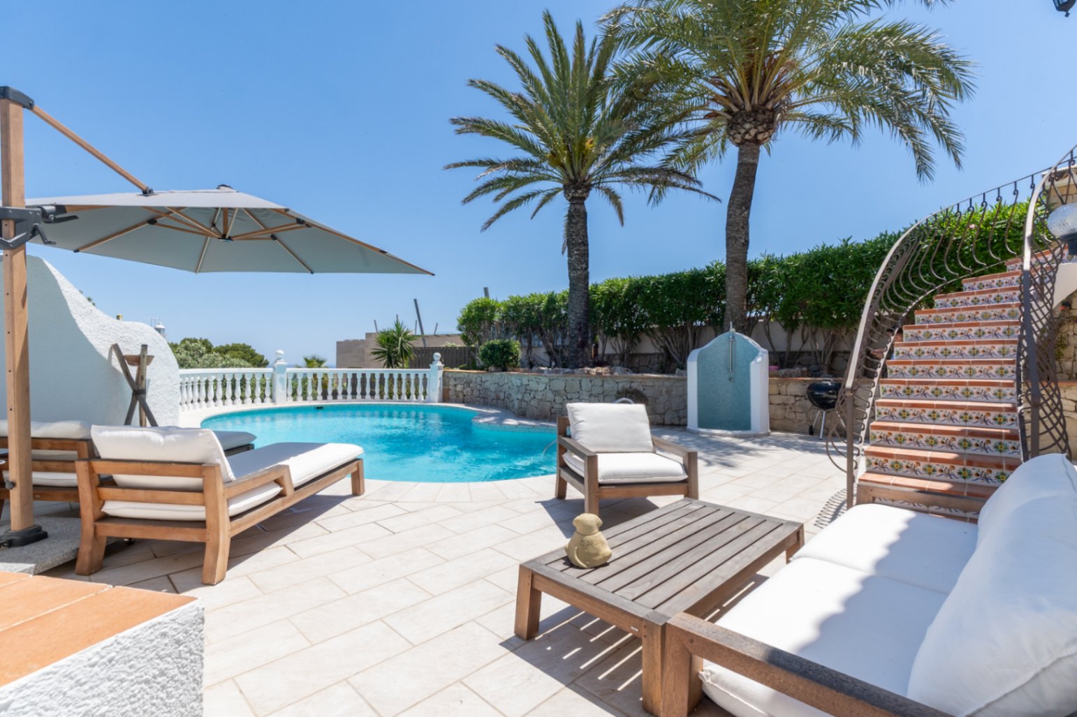 Vaste terrasse de piscine avec chaises longues en bois, un salon de jardin et un escalier courbé aux contremarches carrelées. Des palmiers et une douche extérieure agrémentent l'espace.