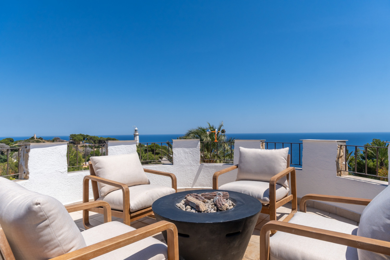 Vaste terrasse sur le toit avec mobilier en bois moderne, foyer central et vues panoramiques sur la mer Méditerranée, incluant un phare lointain.
