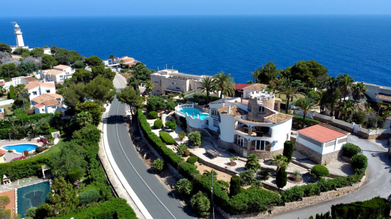 Vue aérienne de la villa méditerranéenne à Jávea, avec piscine surélevée, jardins en terrasses et architecture traditionnelle, face à la vaste mer bleue et un phare lointain.