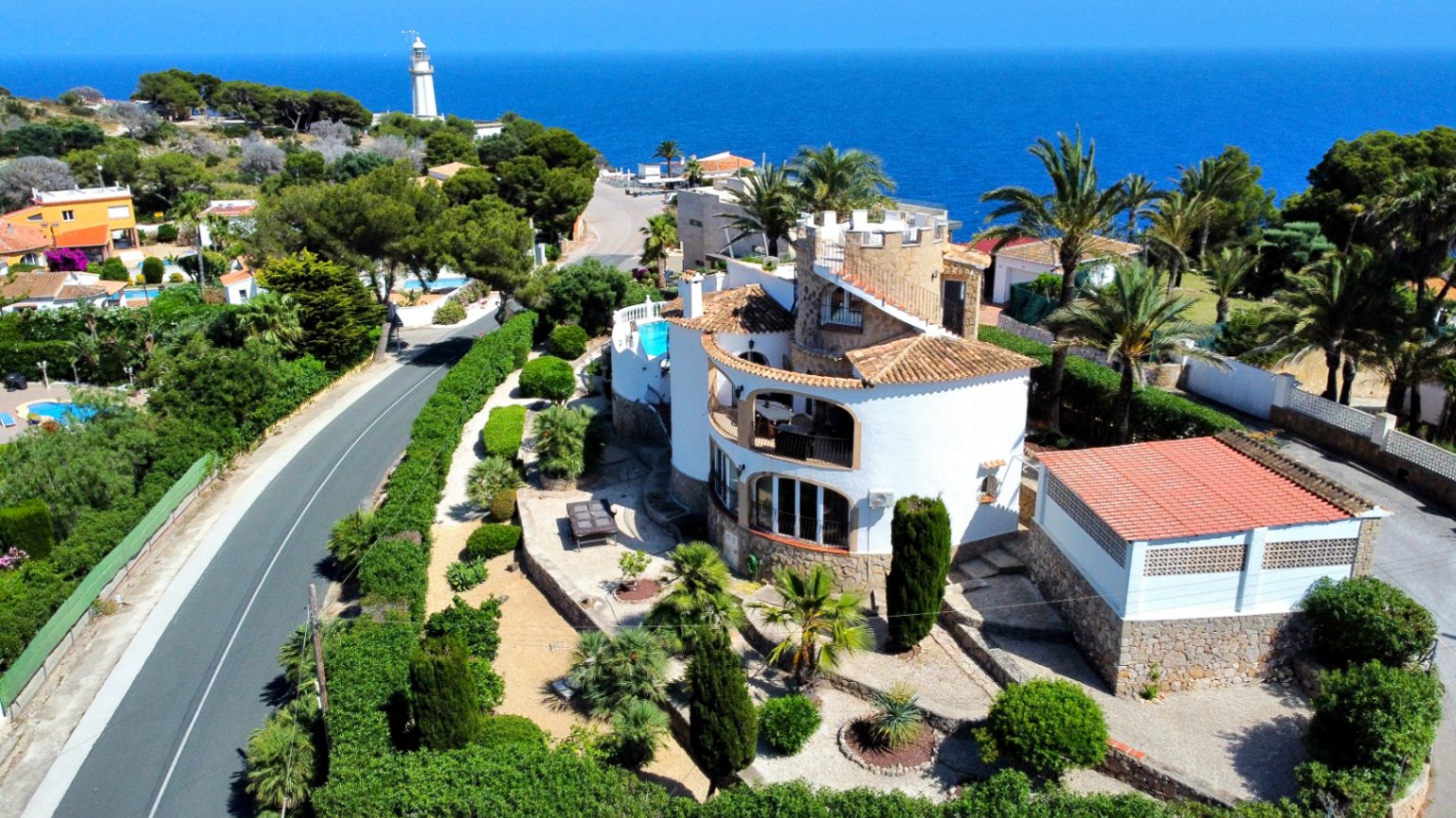 Vue aérienne d'une villa méditerranéenne avec murs blancs, tuiles et piscine. Des jardins luxuriants entourent la propriété, avec la mer et un phare en arrière-plan.