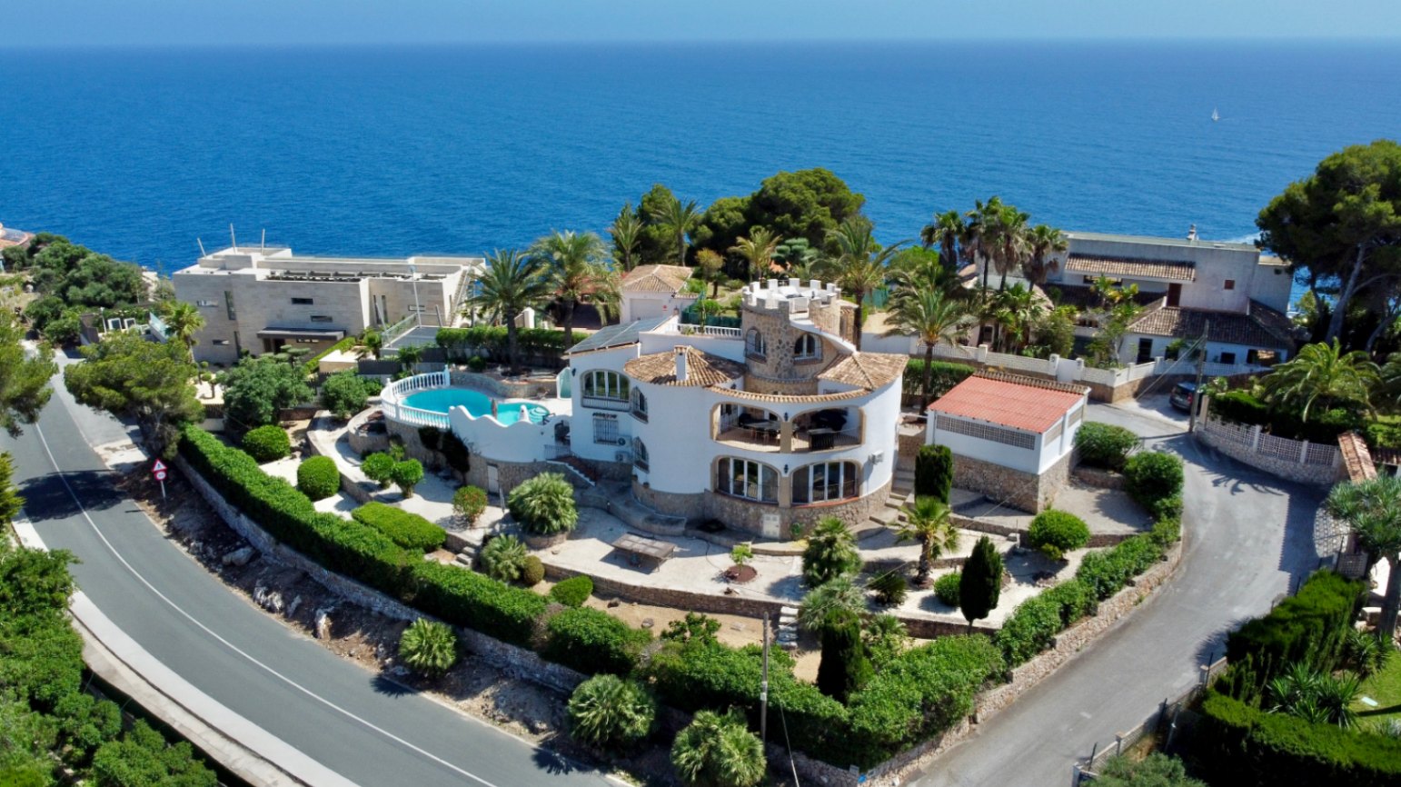 Vue aérienne d'une villa méditerranéenne avec tour ronde distinctive, jardins en terrasses et piscine, le tout face à la vaste mer bleue.