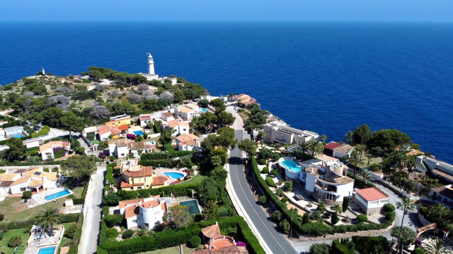 Vue aérienne panoramique d'une urbanisation côtière avec villas, piscines privées et jardins luxuriants, bordant la mer Méditerranée d'un bleu profond et un phare lointain.