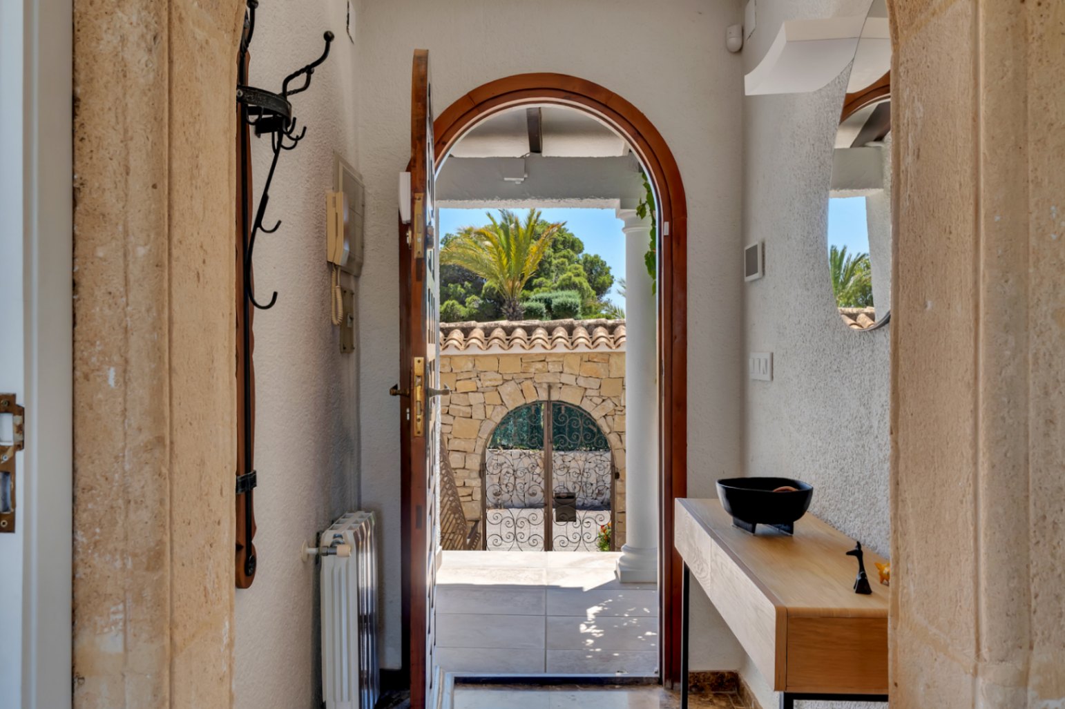 Vue depuis le hall d'entrée à travers une porte en bois cintrée vers un mur de pierre avec portail en fer forgé et végétation méditerranéenne luxuriante. À l'intérieur, console en bois et miroir.