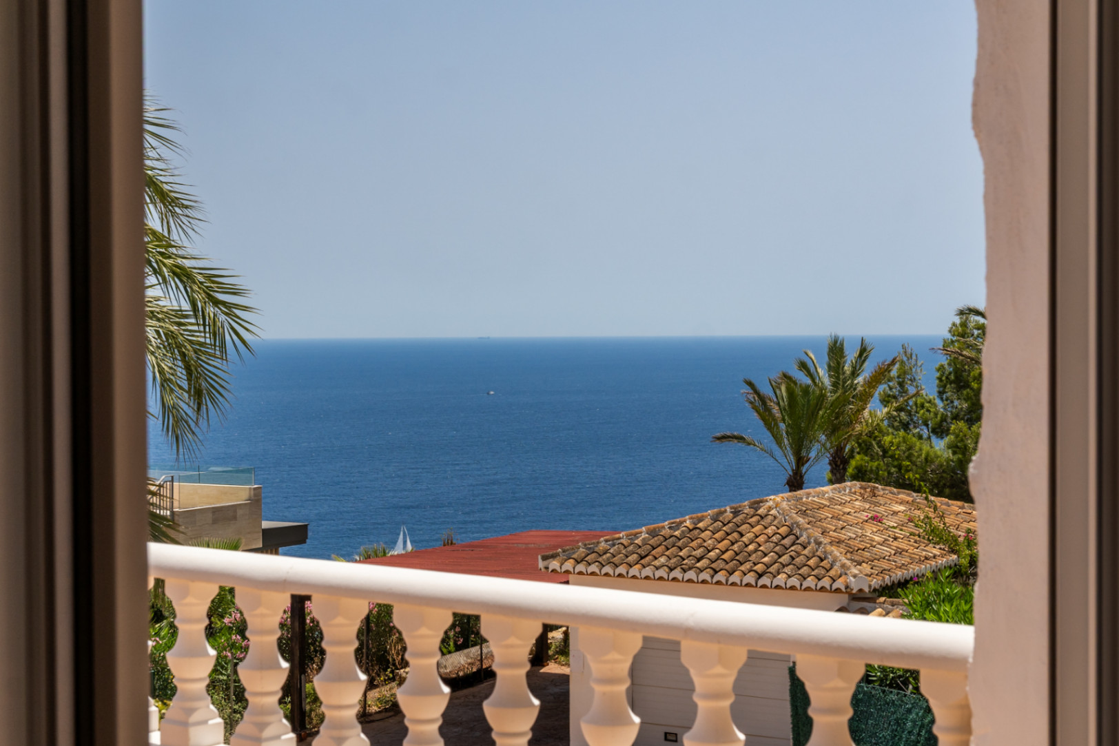 Vue depuis un balcon avec balustrade blanche, toit en terre cuite et palmiers luxuriants, surplombant la vaste mer Méditerranée avec un voilier au loin.