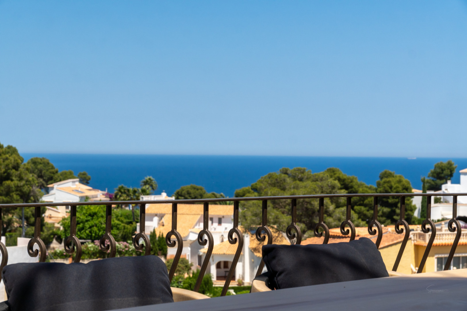 Vue depuis une terrasse avec balustrade en fer forgé, surplombant des toits en terre cuite, des pins et la mer Méditerranée.