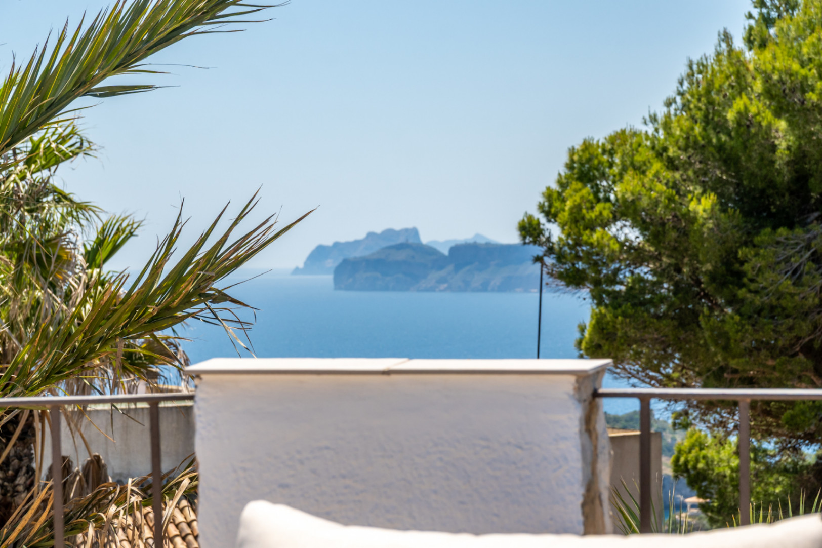 Vue panoramique sur la mer avec une île montagneuse lointaine, encadrée par une végétation méditerranéenne et une balustrade de terrasse.