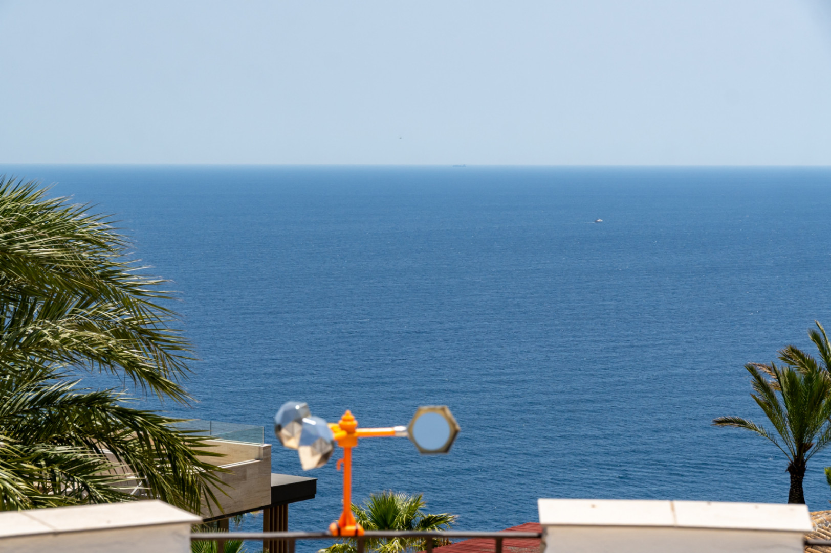 Vue panoramique sur la mer Méditerranée encadrée par des palmiers, avec le bord d'une terrasse moderne et une girouette décorative au premier plan.