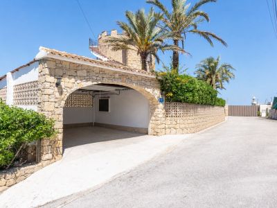 Abri de voiture voûté en pierre offrant un parking couvert, intégré à la villa de style méditerranéen avec escalier en colimaçon menant à une terrasse supérieure. Palmiers et verdure.