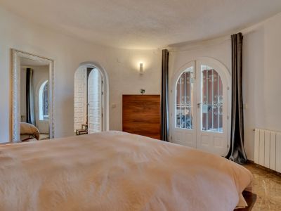 Chambre avec doubles portes cintrées blanches et grilles vers l'extérieur, sol effet marbre et commode en bois. Un grand miroir orné et des murs texturés enrichissent l'espace.