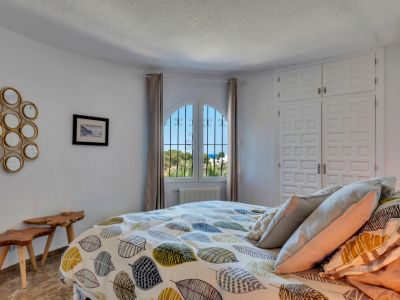 Chambre lumineuse avec fenêtre cintrée et barreaux en fer forgé, offrant une vue partielle sur la mer. Elle dispose d'armoires encastrées blanches et de tables de chevet en bois rustique.