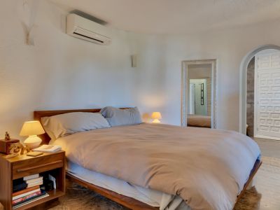 Chambre principale avec lit en bois, murs blancs texturés et climatisation. Un miroir orné et une arche mènent à une porte à motifs distinctifs.