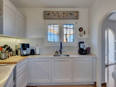 Cuisine moderne avec des armoires blanches, des comptoirs clairs et une crédence en mosaïque. Une fenêtre grillagée donne sur l'extérieur, et une arche mène à un espace lumineux adjacent.